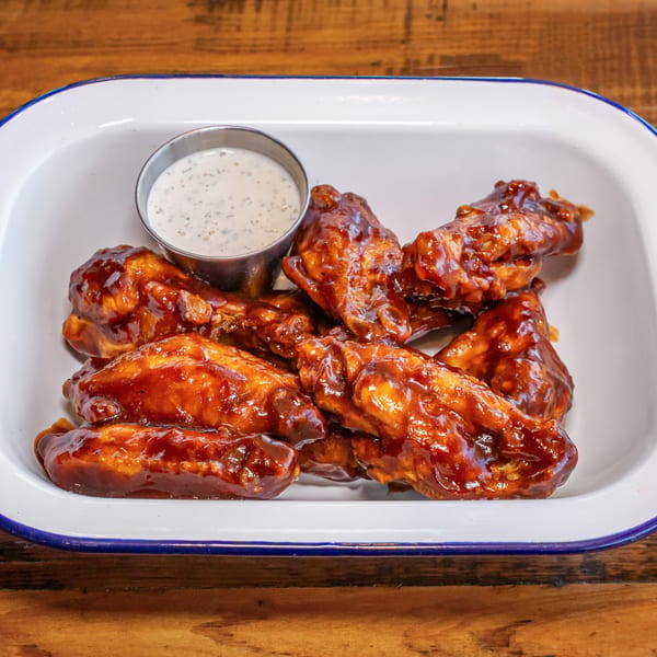 NY Pie's BBQ chicken wings with ranch dressing in a white enamel dish on a wooden table.