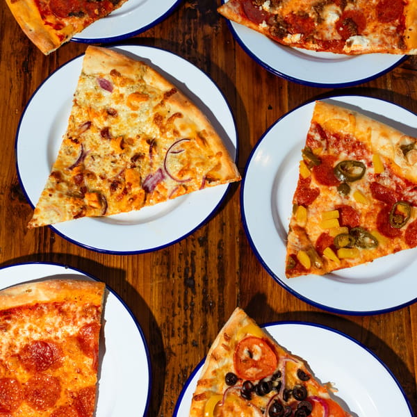 Overhead view of five NY Pie pizza slices with various toppings on white plates.
