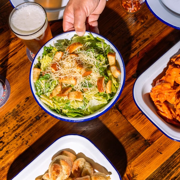 A hand placing a Caesar salad with croutons and parmesan cheese on a wooden table with other dishes.
