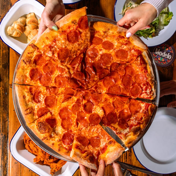 Hands reaching for slices of a large pepperoni pizza on a wooden table with sides.