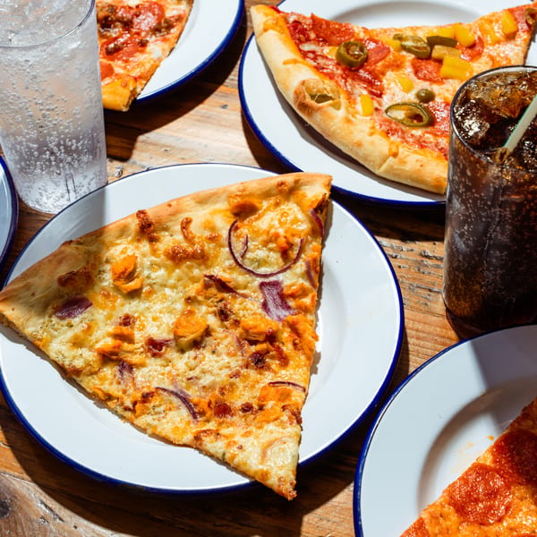 Slices of NY Pie pizza, including pepperoni, chicken, and jalapeño, with drinks on a wooden table.